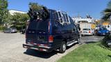 A blue 2013 Ford Econoline van with a roof rack and ladders attached at the back parked in a lot