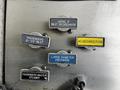 Four metal valve labels on a fire truck panel indicating function, including aerial inlet, passenger inlets, large diameter discharge, and a foam discharge