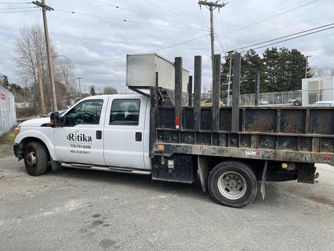 A white 2011 Ford F-350 SD truck with a flatbed and vertical stake pockets on the back displaying a logo on the side