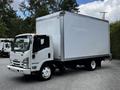 A 2018 Isuzu NPR delivery truck with a white box cargo area and large front windshield parked at an angle with its headlights on