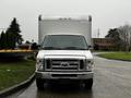 A 2017 Ford Econoline van with a white front and chrome grille parked in front of a large box truck