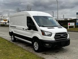 A white 2020 Ford Transit van with a sleek design and black accents is parked on a street facing the viewer. The vehicle features a prominent front grille and alloy wheels