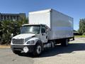 A 2015 Freightliner M2 106 truck with a white box cargo area and a gray front grille is parked at an angle with its headlights on