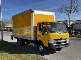 A bright yellow 2017 Hino 155 box truck with a silver roof and a spacious cargo area is parked on a street