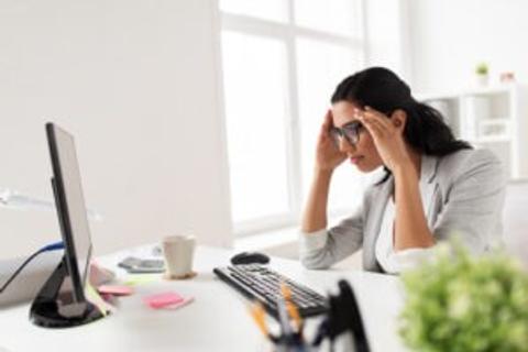 A woman in a blazer sitting at a desk with a computer and a coffee cup appears to be concentrating while holding her temples, indicating focus or stress during a brain training session