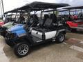 A blue and black golf cart with two seats an open side and large off-road tires parked on a wet surface with other similar vehicles in the background