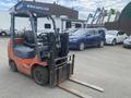 A red and black 2010 Toyota 7FGGU25 forklift with a dual fork attachment positioned in front of parked vehicles
