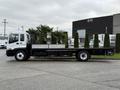 A 2007 GMC F7500 flatbed truck with a white cab and black flatbed, featuring a raised cargo area and surrounded by greenery and palm trees