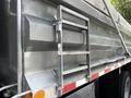 A side view of a metallic dump truck bed with a foldable ladder mounted on the side and a red reflector strip along the bottom