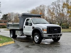 A 2008 Ford F-550 dump truck with a white exterior and gray dump bed parked at an angle with its headlights and turn signals on