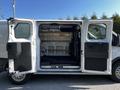 A 2023 Ford Transit van with open double doors revealing a spacious cargo area equipped with shelves and a black rubber mat on the floor