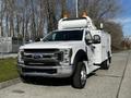 A white 2018 Ford F-550 truck with a utility body and amber lights on the roof parked on a paved surface