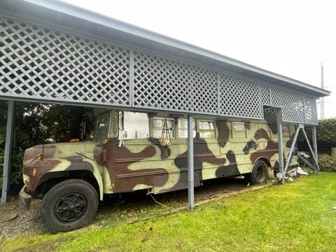 A 1985 Ford B600 painted in camouflage pattern with large black wheels parked under a covered structure