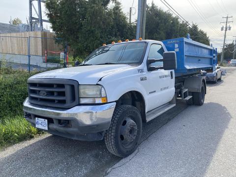 A white 2002 Ford F-550 with orange clearance lights parked beside a blue dump truck. The truck has a flatbed and is equipped for heavy-duty hauling