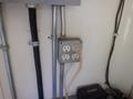 A metal electrical outlet box with three outlets and one plugged cord mounted on a wall with visible wiring and pipes attached