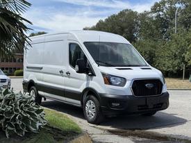 A 2022 Ford Transit van with a white exterior is parked, featuring a black front grille and headlights, and has a high roof design and sliding side doors