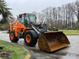 A 2018 Doosan DL300-5 wheeled loader with a large bucket in the front and rugged tires is parked on a paved surface with greenery nearby