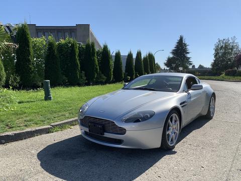 A silver 2008 Aston Martin Vantage with a sleek design featuring a low profile and distinctive grille parked on a gravel road.