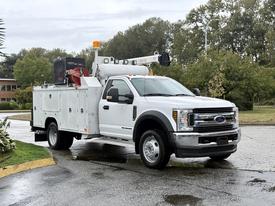 A 2018 Ford F-550 utility truck with a white service body and mounted equipment on the back, featuring a raised boom and orange safety light