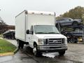 A 2013 Ford Econoline box truck with a white cargo area parked in a lot with additional vehicles on a trailer in the background