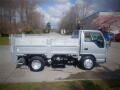 A silver 2008 Isuzu Elf flatbed truck parked on a street with a low cab and a flatbed cargo area featuring foldable sides