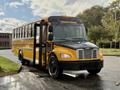 A yellow and black 2022 Freightliner B2 school bus with front doors open is parked on a wet surface with trees in the background