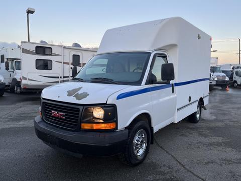 A 2009 GMC Savana van with a white body and a blue stripe on the side featuring a box-shaped cargo area and visible wear on the front hood