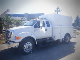 A white 2005 Ford F-750 truck with a large utility box and equipment compartments on the back parked on the street