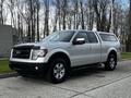 A silver 2014 Ford F-150 truck with a cap on the bed parked on a concrete surface facing left