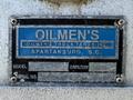 A blue and black metal nameplate displaying the text "OILMEN'S OILMEN'S TRUCK TANKING INC SPARTANBURG SC MODEL CAPACITY SERIAL NO" with visible wear and scratches