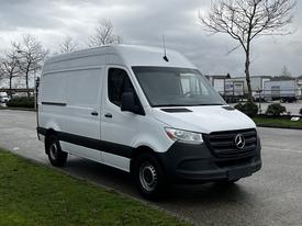 2021 Mercedes-Benz Sprinter van in white with a black front grille parked on a street with green grass and trees nearby