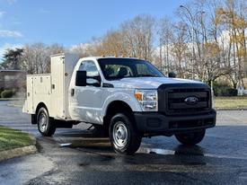 A white 2012 Ford F-350 with a black front grille and utility box in the cargo area, featuring orange marker lights on the roof and a robust truck design