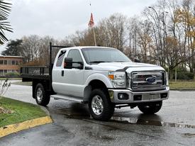 A 2016 Ford F-350 SD pickup truck with a flatbed and a black metal railing stands on a wet surface with trees in the background