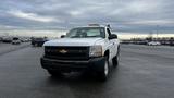 A 2013 Chevrolet Silverado 1500 with a white exterior and a black grille is parked in an open area with its headlights on and a yellow light bar on the roof