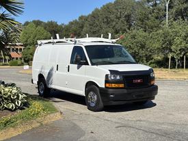 A white 2019 GMC Savana van with a ladder rack on the roof parked on a street