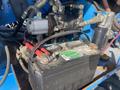 A close-up view of a battery with red and black cables connected, located in a machinery engine compartment with various metal components and hoses visible around it