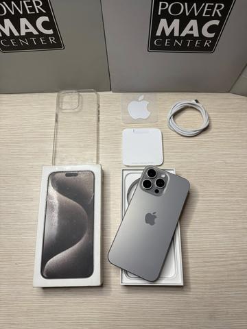 An iPhone 15 Pro Max in silver positioned on its packaging with a clear case wireless charging pad and white charging cable arranged nearby