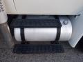 A silver fuel tank with black straps and step treads underneath a truck's chassis