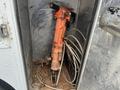 A large orange jackhammer leaning against a wall inside a trailer with some coiled hoses and chains visible nearby