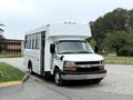 A white 2015 Chevrolet Express shuttle bus with a high roof and large side windows parked at an angle with the front facing the viewer