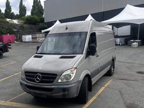 A 2010 Mercedes-Benz Sprinter van in a light gray color with a high roof and sliding side door positioned in a parking area
