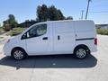 A white 2019 Nissan NV200 cargo van parked on a gravel surface with a sliding side door and standard wheels