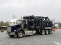 A 2018 Kenworth T800 truck with a black and silver exterior, featuring a large equipment attachment in the back, positioned on gravel ground