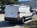 A white 2016 Ford Transit van with a roof rack and sliding doors parked in a lot