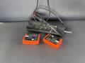 A black device charger with two orange battery packs positioned beside it and a black wire connected to the charger