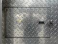 A metal access door with a diamond plate texture featuring a hinge and a latch labeled Outtrans Obstructions