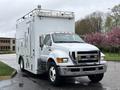 A white 2011 Ford F-650 with a large boxy rear compartment and a raised roof equipped with lights and a ladder on top