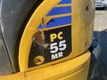 A close-up of a yellow and black Komatsu PC55MR2 excavator section showing a damaged logo with peeling paint and exposed metal