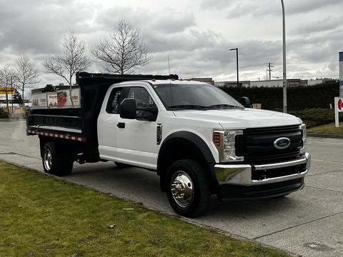 A 2019 Ford F-550 with a white exterior and a dump bed is parked on a grassy area featuring large wheels and a prominent front grille