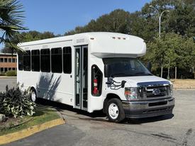 A white 2021 Ford Econoline bus with large windows and an open door sits on a paved surface showcasing its spacious cabin and passenger access features